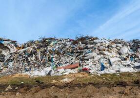 A large mound of garbage is visible at a landfill, consisting of various waste types including plastic bags and discarded furniture, illuminated by sunlight on a clear day photo