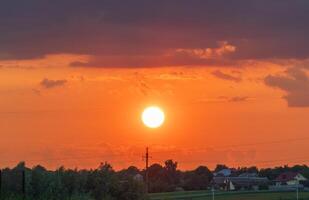 Vibrant sunset casts an orange glow over a rural landscape. Silhouettes of houses and trees create a peaceful atmosphere as day transitions into night photo
