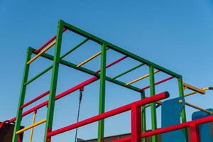 A colorful playground structure stands tall under a clear blue sky. The vibrant red, green, and yellow bars invite children to climb and play in the afternoon sun photo