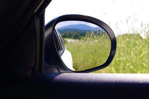 A side mirror reflects a green field and forested hills under a bright sky, captured from inside a car to convey a peaceful travel moment in nature. photo