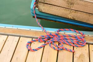 A colorful twisted red and blue rope lies loosely on a wooden dock near calm green water. The rope is attached to a wooden boat, creating a nautical mood. photo
