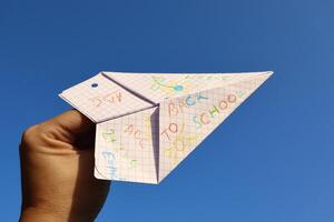 Paper airplane made from a school notebook, held in hand against a clear blue sky. Colorful drawings and text say BACK TO SCHOOL symbolizing learning. photo