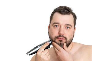 A young man with casual clothing is using an electric shaver to groom and shave his well-maintained beard. The image showcases personal grooming and hygiene against a clean white background. photo