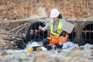Environmental Engineer Testing Water Quality in Urban Drainage, Scientist Collecting Water Samples from Industrial Runoff, Female Technician Monitoring Water Pollution Near Concrete Pipes photo