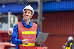 Engineer man with laptop inspecting containers box at shipping port, Foreman worker with blurred female engineer in background working at shipping yard photo