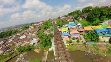 Dynamic FPV drone flythrough over railway bridge and colorful village in Malang, Indonesia, showcasing vibrant rainbow houses, riverside houses, and tropical local life from above. video