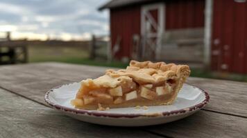 A slice of a pple pie on a plate sitting on a wooden table with a barn in the background outdoors photo