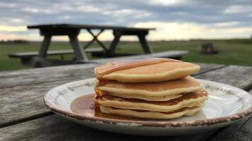 Stack of pancakes with syrup on a plate sitting on a wooden table outdoors with a picnic table behind photo