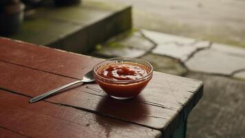 A bowl of ketchup with a spoon on a rustic wooden table near a blurred outdoor background scene view photo