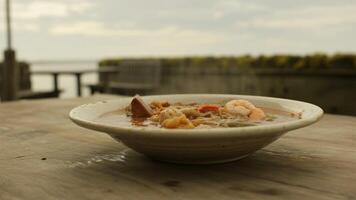 A bowl of steaming soup with shrimp and other ingredients on a wooden table near the ocean view photo