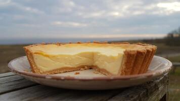 A partially eaten custard tart on a plate with a sea and cloudy sky in the background outside view photo