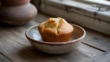 A single muffin in a bowl on a wooden surface near a window with a clay pot in the background photo