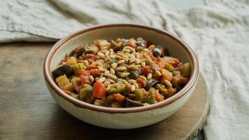 A bowl of ratatouille topped with pine nuts on a wooden board with a linen cloth background view photo