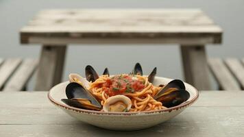 A bowl of spaghetti with mussels and clams on a white table with a bench in the background photo