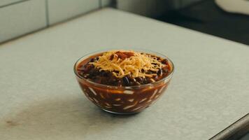 A clear bowl filled with chili and topped with shredded cheese sitting on a white surface top photo