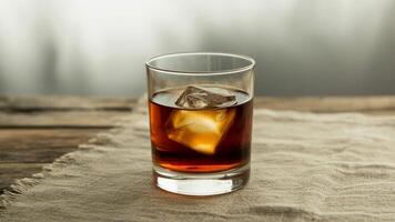 A glass of whiskey with a large ice cube on a wooden surface with a textured cloth underneath it shown photo