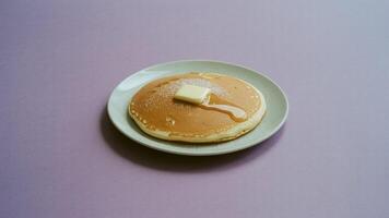 A single pancake on a plate with butter and syrup against a light purple background in a studio shot photo