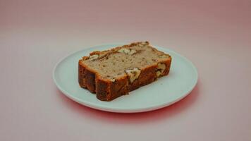 A slice of banana nut bread on a white plate against a light pink background in a studio setting photo