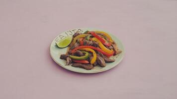 A plate of fajitas with beef and bell peppers and a lime wedge on a light pink background color photo
