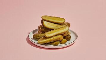 A stack of sliced pickles on a white plate against a light pink background in a studio setting photo