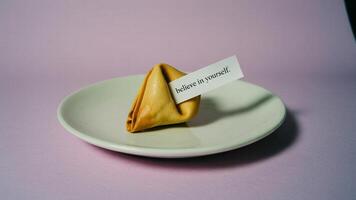 A fortune cookie with a message that says believe in yourself on a white plate with pink background photo