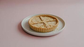 A tart with a patterned crust sits on a white plate against a light pink background in a studio shot photo