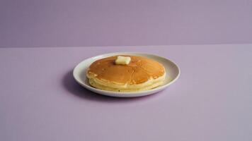 Stack of pancakes topped with butter on a white plate against a lavender background in a studio shot photo
