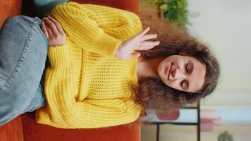 Young child girl smiling friendly at camera and waving hands gesturing hello, hi, greeting or goodbye, welcoming with hospitable expression. Teenager kid at home in living room sits on sofa. Vertical video