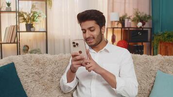 Indian young man at home sitting on sofa focused on smartphone typing text, browsing news, reacting to notifications. Arabian guy using mobile phone for social media, work, texting, digital lifestyle video