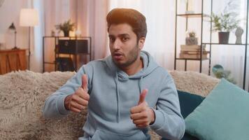 Happy cheerful Indian man looking approvingly at camera, showing ok gesture, positive like sign. Arabian Hindu guy sitting on sofa in modern apartment, smiling confidently, approving something good. video