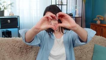 Cheerful young woman at home sitting on sofa showing heart gesture, happily expressing positive emotions. Caucasian girl sending love, friendship, joy, warm feelings, pleasant emotional attitude video