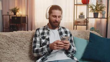 Young man sits at home with wireless headphones, scrolling phone feed and listening to music. Guy on sofa removes earphones for an incoming call, starting a lively, fun cheerful conversation talk video