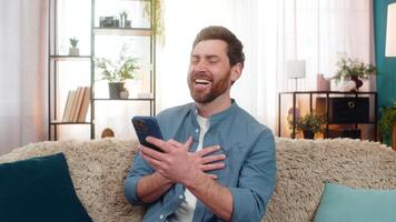 Young man at home laughing while reading funny social media comments on smartphone. Happy Caucasian guy sitting on sofa, holding mobile phone, enjoying entertaining videos online content interactions