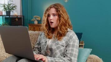 Young woman at home sitting with laptop suddenly raising eyebrows surprised by unexpected positive news. Caucasian redhead girl on sofa reacts with curiosity and joy to interesting discovery online video