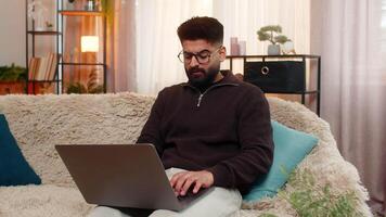 Indian man at home types on laptop with focused expression, preparing for upcoming online exam with full attention. Arabian guy on sofa concentrates on studying and memorizing materials for success video