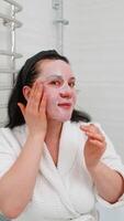Middle-aged mature woman carefully applies fabric sheet mask, gently smoothing edges around face. Lady thoughtfully adjusts mask with fingers, checking reflection in mirror with calm expression. POV video