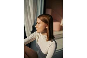 Woman in white turtleneck sitting near window with sunlight casting shadows indoors photo
