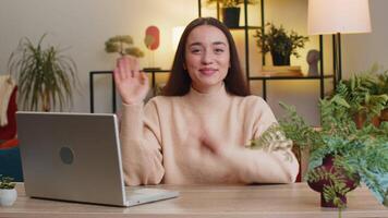 Hello. Pretty young woman smiling friendly at camera, waving hands gesturing hello hi, greeting or goodbye welcoming with hospitable expression at home office. Caucasian girl sitting at table in room video