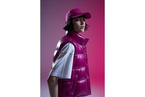 A woman with short hair and a pink cap wearing a puffy vest in front of a pink wall photo