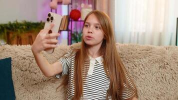 Young child girl sits on sofa happily receives call from friend on smartphone, waves hand with joy. School kid smiles, talks cheerfully, enjoying friendly conversation at home with mobile device video