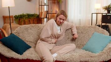 Young adult woman holds her stomach while sits on home sofa reacting to sharp pain from overeating or cramps. Blonde girl expresses visible discomfort showing emotional strain from digestive problem video