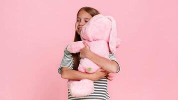Young child girl gently hugs a plush toy bunny with love and happiness, smiling sweetly and closing eyes in joy, expressing warmth and emotional connection. School kid isolated on pink background. video