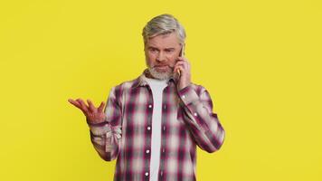 Mature man speaks on mobile phone with forceful tone, revealing anger agitation during tense talk quarrel. Middle-aged guy on yellow background shows argumentative stance, emphasizing heated dispute. video
