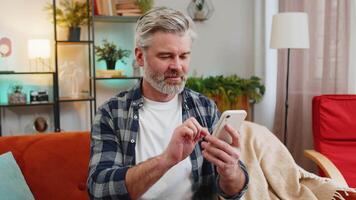 Happy mature man sitting on sofa at home getting message from old friend using smartphone look aside with smile. Caucasian grey-haired old man reading good news on phone screen in living room at home. video