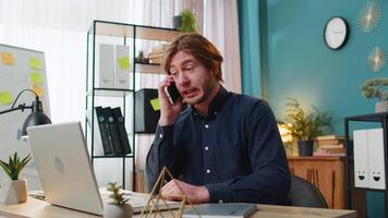 Young businessman freelancer answering to client on mobile phone call, using smartphone and laptop. Conversation. Caucasian man guy working online remote distance job, talking at home office desk. video