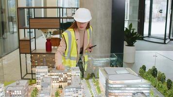 Confident female girl architect in hardhat and reflective jacket using smartphone and analyzing business office complex architectural model. Woman lady smart designer of buildings working in office. video