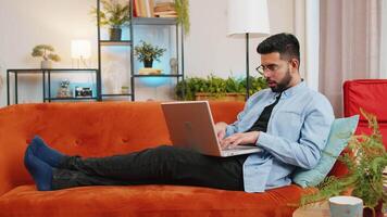Full length of smiling Indian man typing on laptop using apps lying on comfortable sofa. Happy young Hispanic guy working on computer in living room at home. E-learning, browsing internet on netbook video