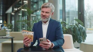 Successful happy rich mature businessman holding smartphone and counting euro bill money fan sitting in office lobby. Happy Caucasian bearded guy winning online lottery game. Calculating budget. video