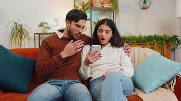 Disappointed multiethnic couple reading together bad news on smartphone while sitting on sofa in room at home. Sad young family are depressed by unpleasant message, shocked by information about loss video