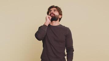 Thoughtful clever handsome Indian man in long sleeves looking aside with positive expression pondering a solution doubting question. Hispanic adult guy with beige studio background. People lifestyles. video